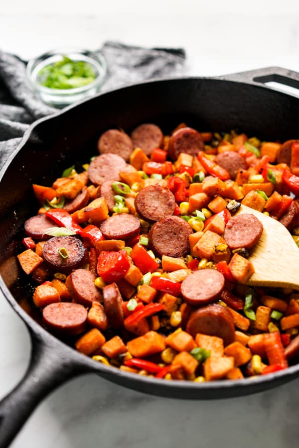 A large cast iron skillet with sweet potatoes, sausage and red bell peppers in it, along with a wooden spatula on the side