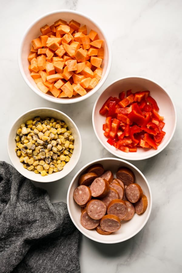 A bowl of diced sweet potatoes, a bowl of sliced red bell peppers, a bowl of sliced kielbasa sausage and a bowl of frozen corn.