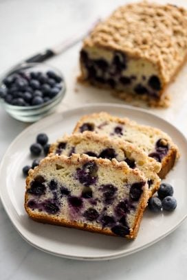Three slices of blueberry bread with crumb topping on a plate with the loaf in the background