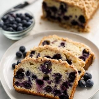 Three slices of blueberry bread with crumb topping on a plate with the loaf in the background