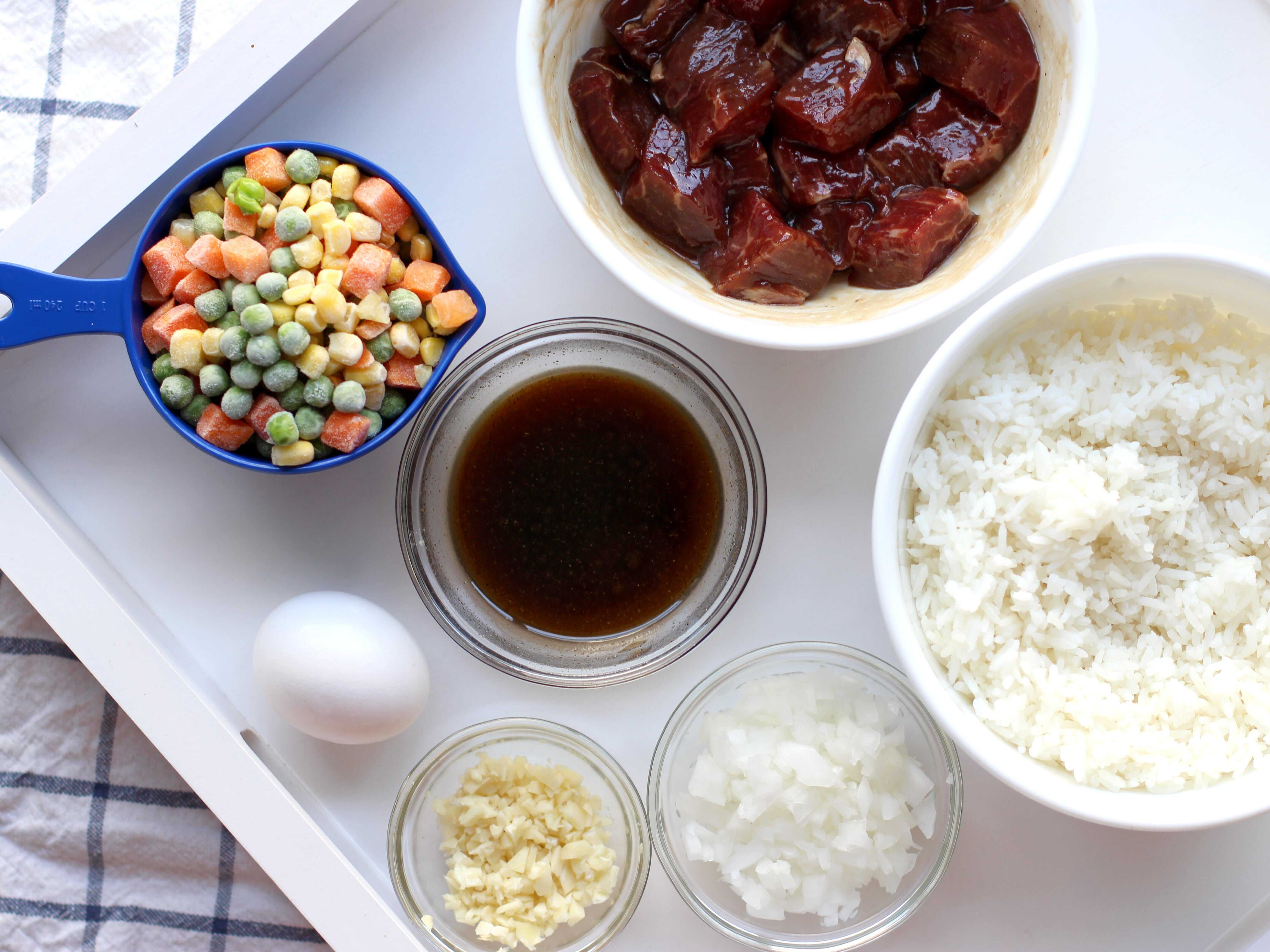 A bowl of white rice, a bowl of marinated steak, mixed vegetables, sauce, garlic, onions and an egg