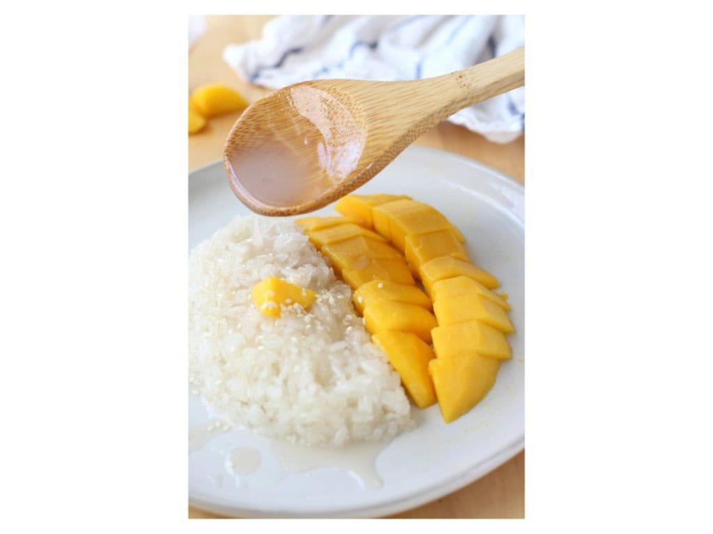 Using a wooden spoon to drizzle sweet coconut sauce onto sticky rice topped with sliced mangoes