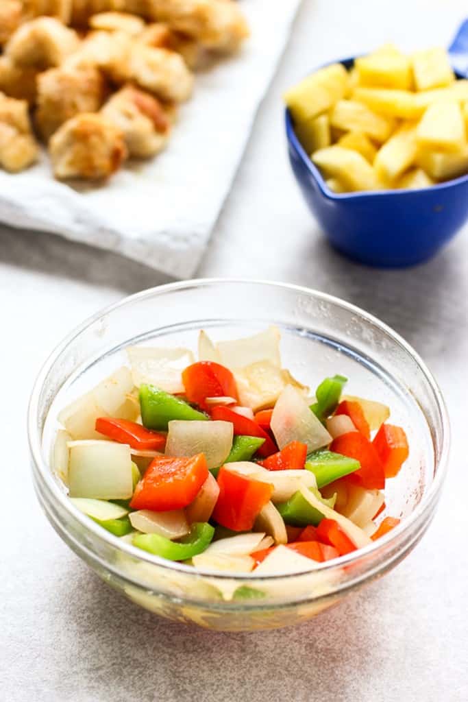 A small bowl of sweet onions and bell peppers, pineapple and chicken in the background