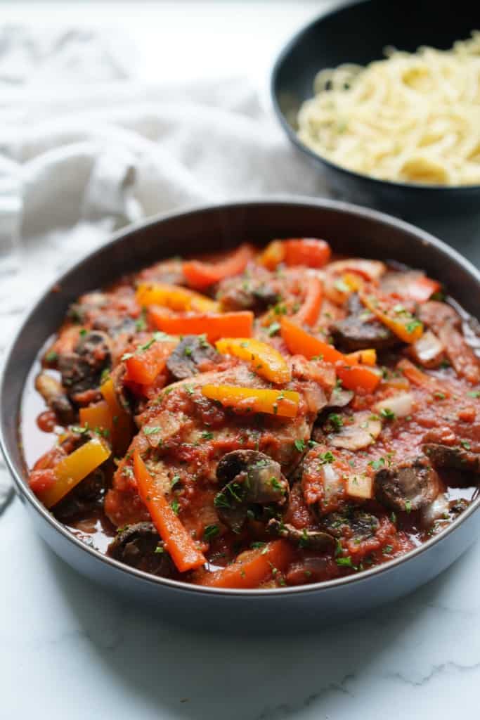 A bowl of chicken cacciatore with a bowl of pasta in the background