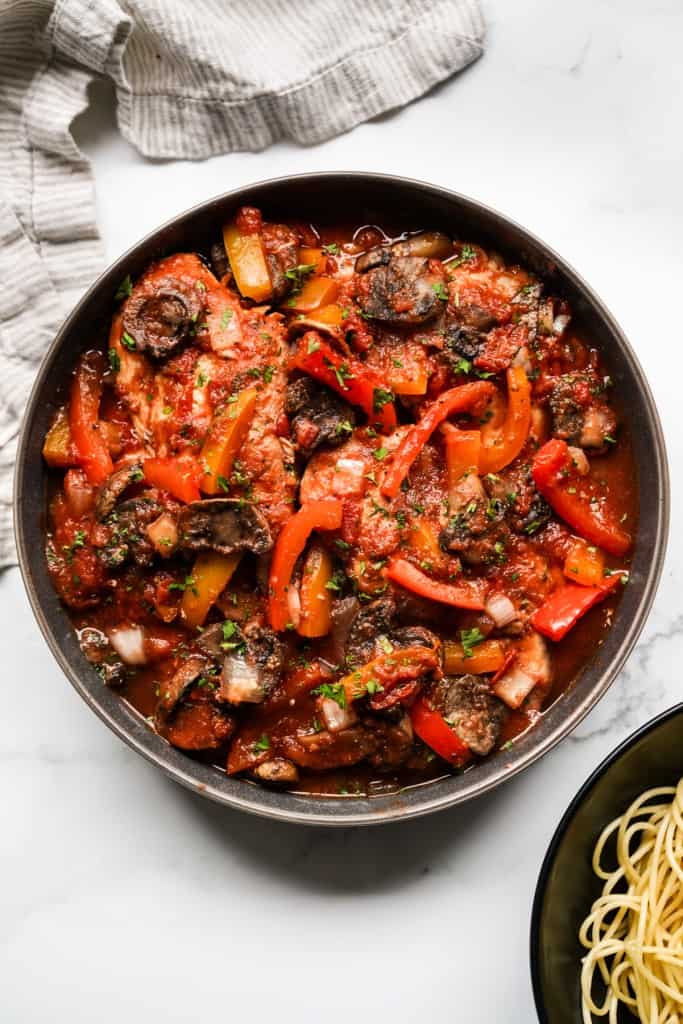 A bowl of chicken cacciatore topped with tomatoes, bell peppers and mushrooms. Spaghetti on the side