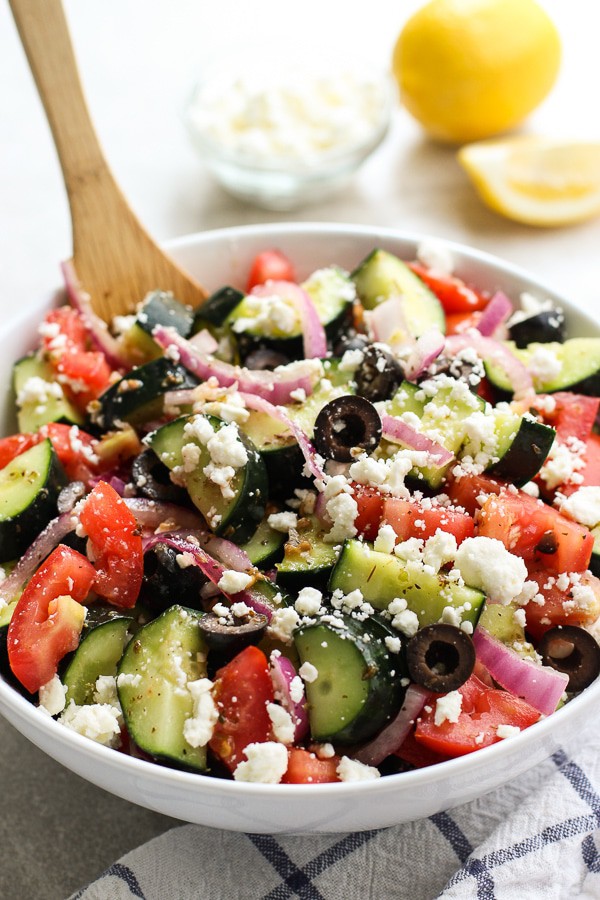 A large bowl of cucumber, tomatoes, red onions and olives tossed in Greek Lemon dressing. Serving spoon in the bowl.