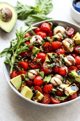 A bowl of caprese salad with avocado with basil and avocado in the background