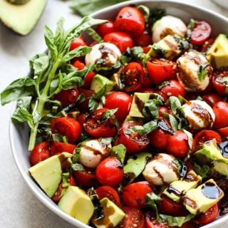 A bowl of caprese salad with avocado with basil and avocado in the background