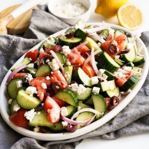 An oval platter of greek cucumber salad tossed with chunky tomatoes, olives, red onions and feta cheese.