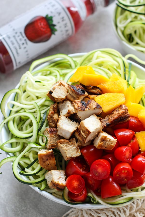 A bowl of zoodles topped with grilled chicken, mango and tomatoes with BRIANNAS dressing on the side