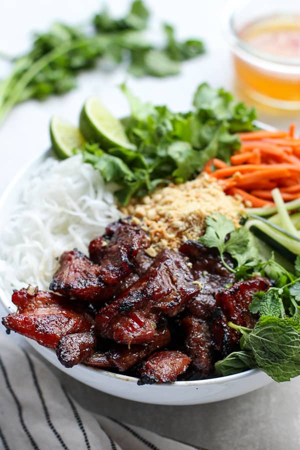 A bowl filled with vermicelli noodles, grilled pork and veggies, with sauce in the background