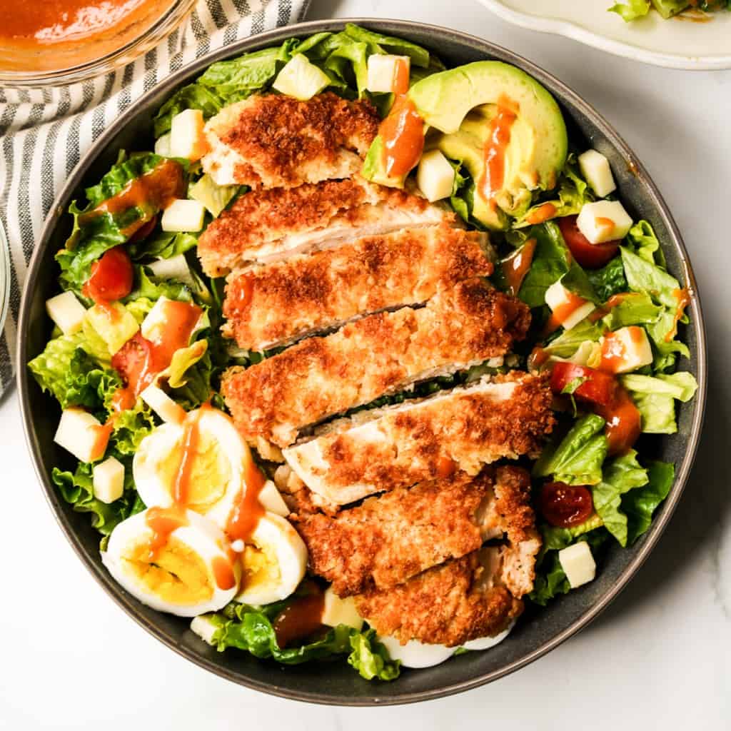 A bowl of crispy chicken on top of a bed of salad with avocado and boiled eggs, drizzled with dressing