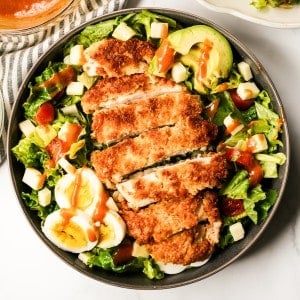 A bowl of crispy chicken on top of a bed of salad with avocado and boiled eggs, drizzled with dressing