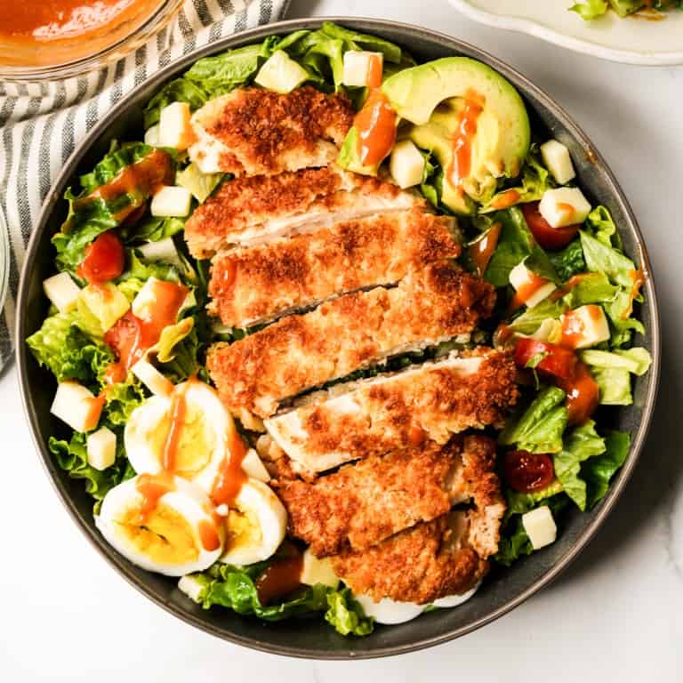 A bowl of crispy chicken on top of a bed of salad with avocado and boiled eggs, drizzled with dressing