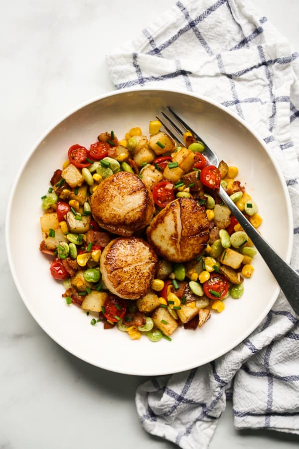 A bowl of corn, lima beans and potatoes topped with seared scallops, and a fork in the bowl.