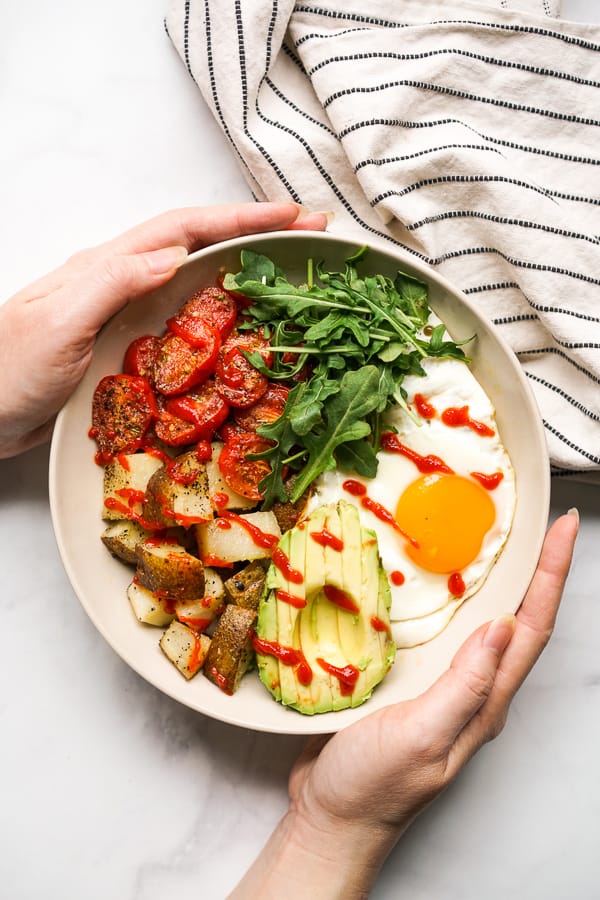 Hands holding a breakfast bowl filled with roasted potatoes, roasted tomatoes, avocados, egg and arugula