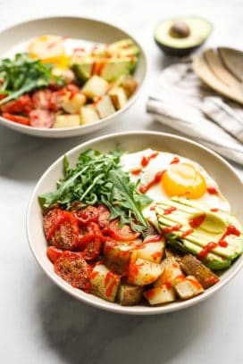 Two savory breakfast bowls filled with roasted potatoes, roasted tomatoes, avocados, egg and arugula