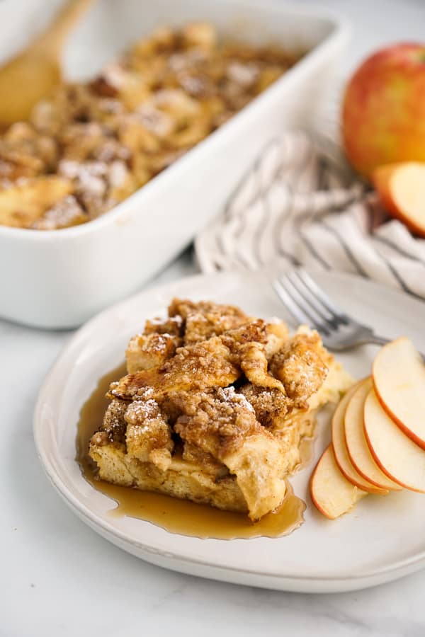 A plate of Apple French Toast Bake with maple syrup on it, apples and a fork on the side