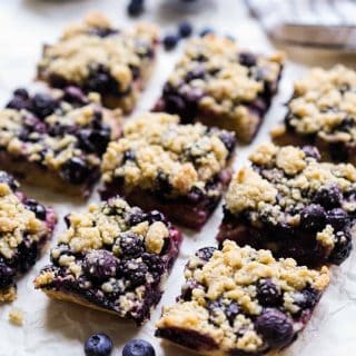 Square blueberry crumble bars on parchment paper with blueberries around it