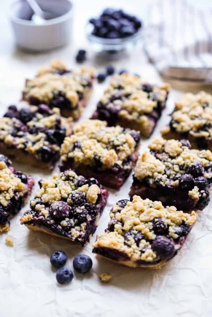 Square blueberry crumble bars on parchment paper with blueberries around it