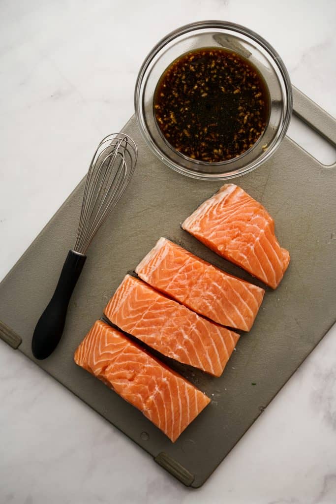 Pieces of raw salmon and a bowl of teriyaki sauce on cutting board