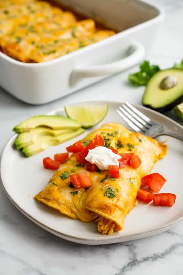 A plate filled with two chicken enchilada, with a casserole of chicken enchiladas in the background