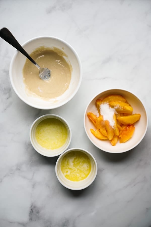 A bowl of cobbler flour topping, a bowl of sliced peaches with sugar, and two ramekins with melted butter