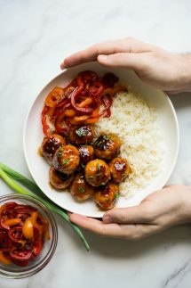 Two hands holding a bowl of sweet and sour meatballs, rice and bell peppers