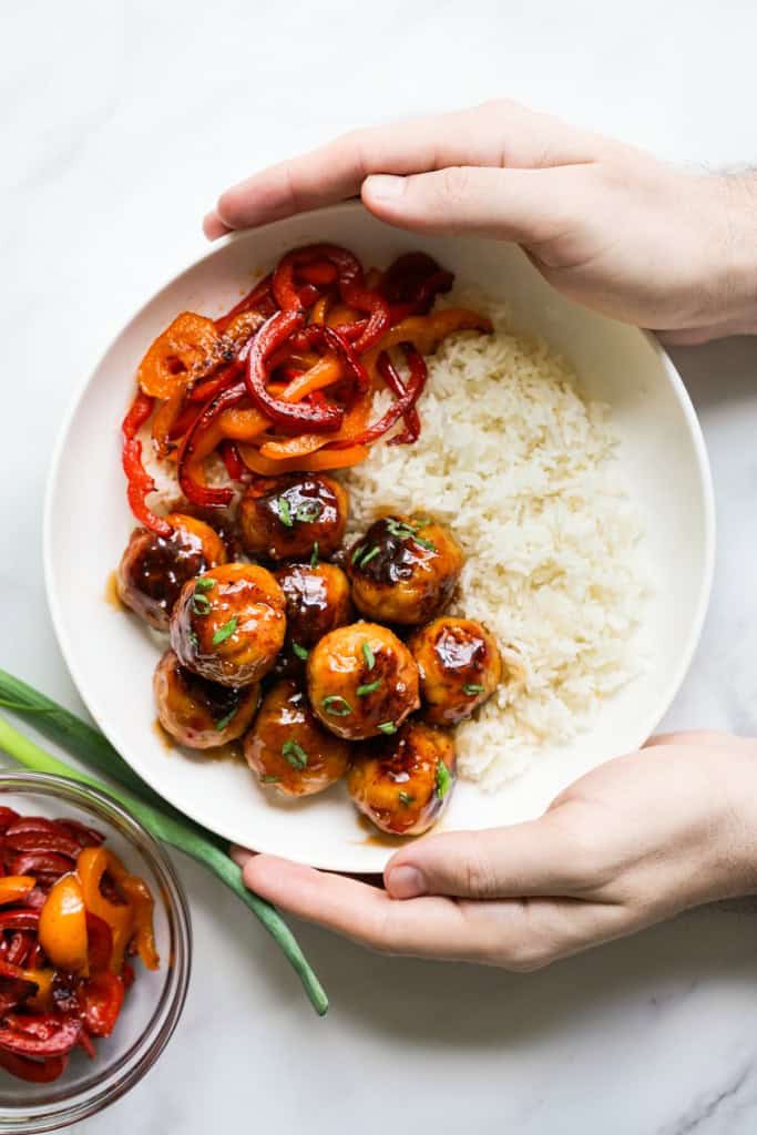 Hands holding a bowl of rice topped with red bell peppers and sweet and sour meatballs
