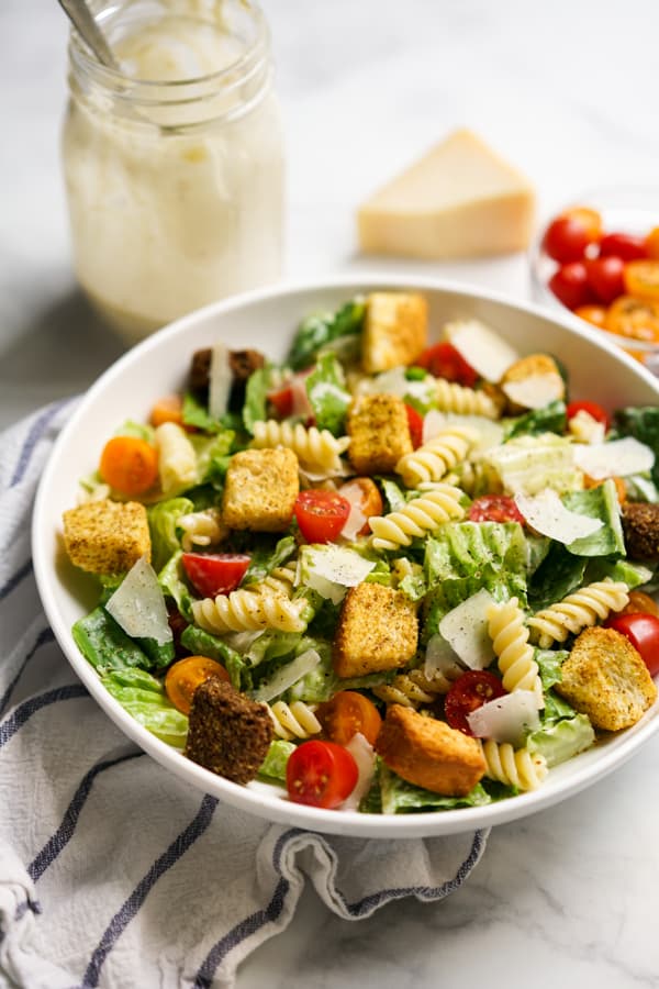 A bowl of Caesar salad with pasta, tomatoes and croutons. Caesar dressing in the background