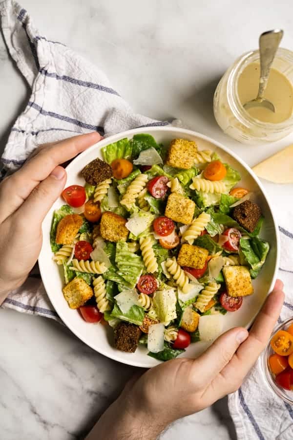 Hands holding a bowl of Caesar Pasta Salad with a mason jar of dressing on the side