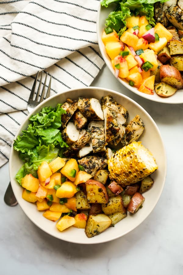 Top down closeup of a bowl filled with grilled chicken, grilled potatoes, lettuce, corn on the cob and peach salsa