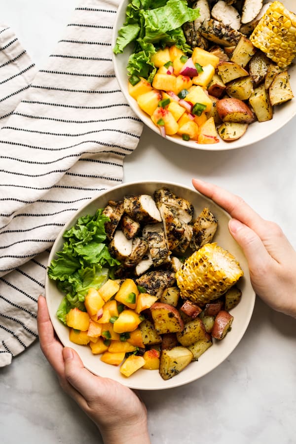 Hands holding a bowl filled with grilled chicken, grilled red potatoes, corn, lettuce and peach salsa