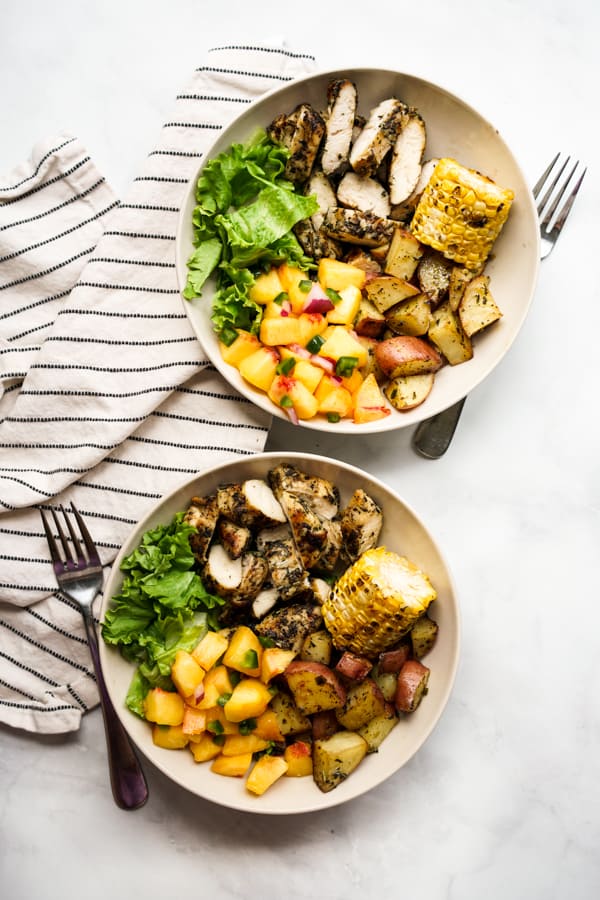 Two bowls of grilled chicken, grilled potatoes, corn on the cob, lettuce and peach salsa, with forks next to each bowl