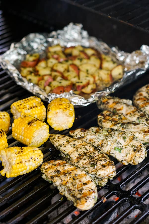 Grilled chicken tenderloins and grilled corn placed directly on the charcoal grill while cubed red potatoes are placed on a foil on the grill