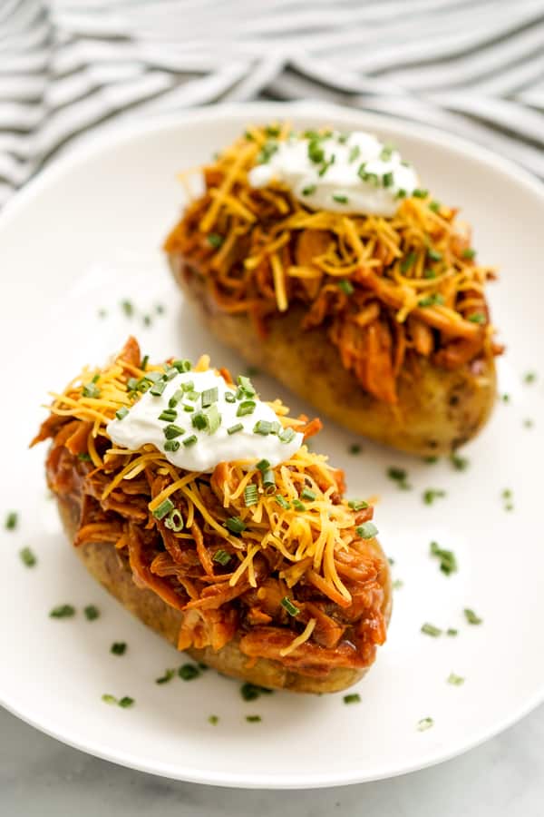 Two baked russet potatoes topped with shredded BBQ chicken, shredded cheddar cheese, sour cream, and chives on a oval plate
