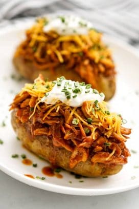 Two baked russet potatoes topped with shredded BBQ chicken, shredded cheddar cheese, sour cream, and chives