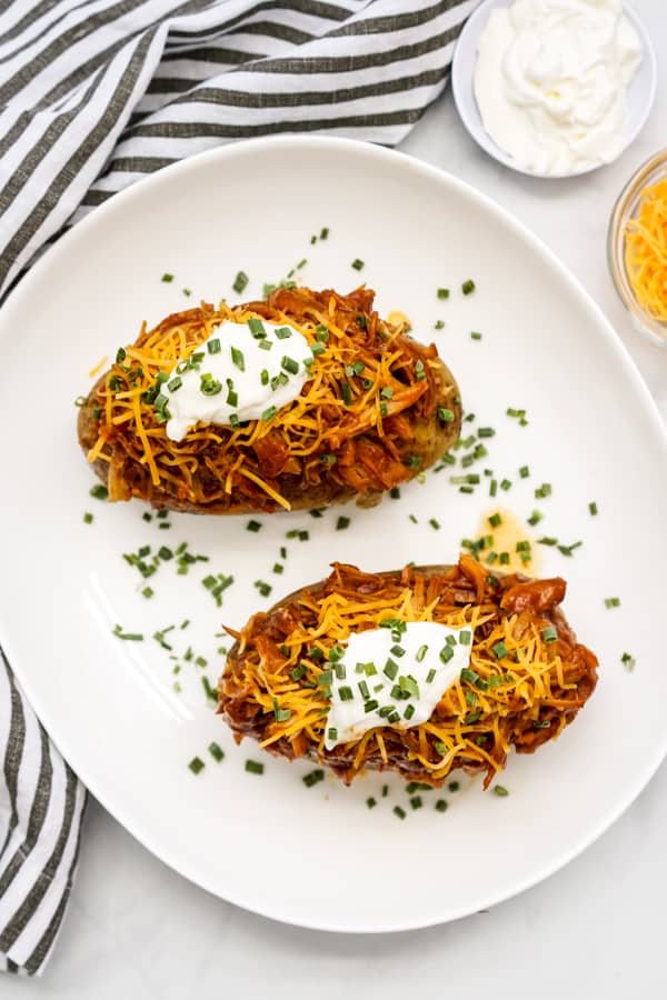 Two baked russet potatoes topped with shredded BBQ chicken, shredded cheddar cheese, sour cream, and chives