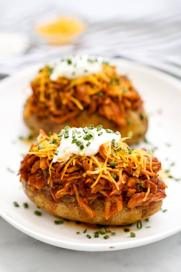 Two baked russet potatoes topped with shredded BBQ chicken, shredded cheddar cheese, sour cream, and chives with a ramekin of shredded cheese in the background