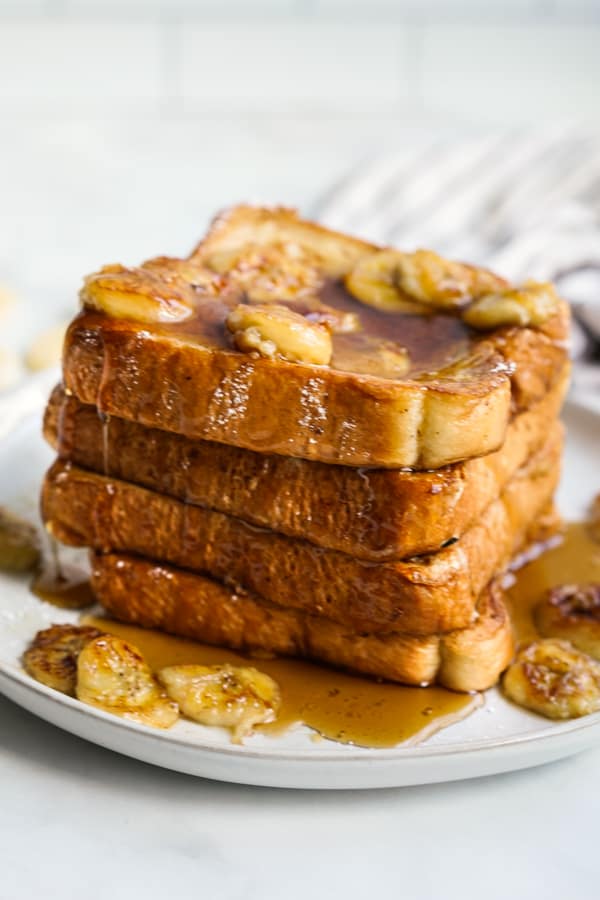 French toast stacked on a plate topped with bananas and maple syrup