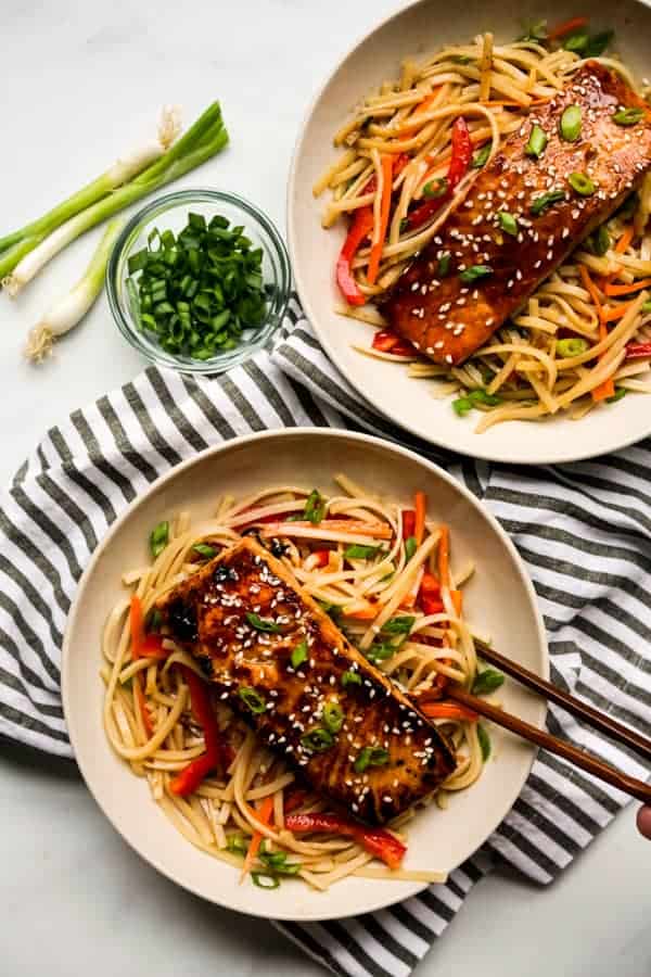 Chopsticks digging into a bowl of noodles topped with orange ginger salmon, another similar bowl also in sight