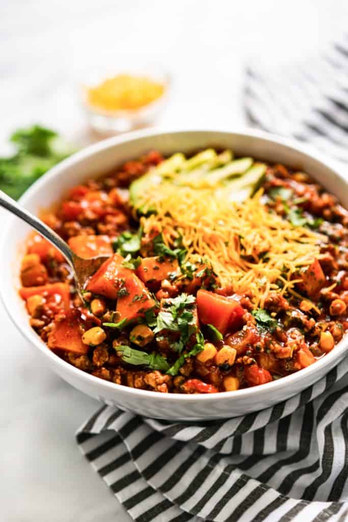 Stirring a bowl of turkey sweet potato chili topped shredded cheese, avocados and cilantro