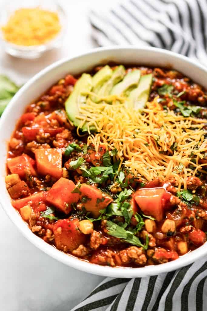 A bowl of sweet potato turkey chili with toppings such as shredded cheese, chopped cilantro and sliced avocados