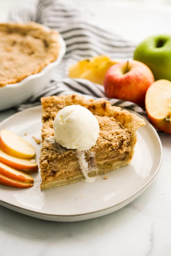 A slice of apple crumble pie topped with vanilla ice cream with a whole pie in the background