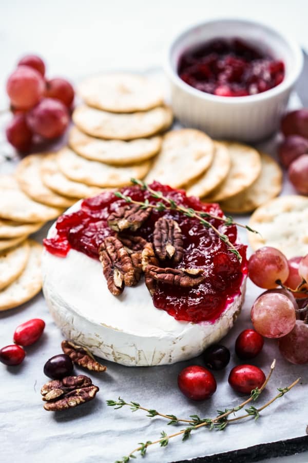 A platter filled with brie topped with cranberry sauce and pecans, crackers and grapes in the background