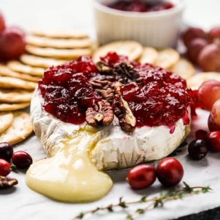 A platter with melted cheese oozing out of baked brie, topped with cranberries and pecans, with crackers surrounding the brie