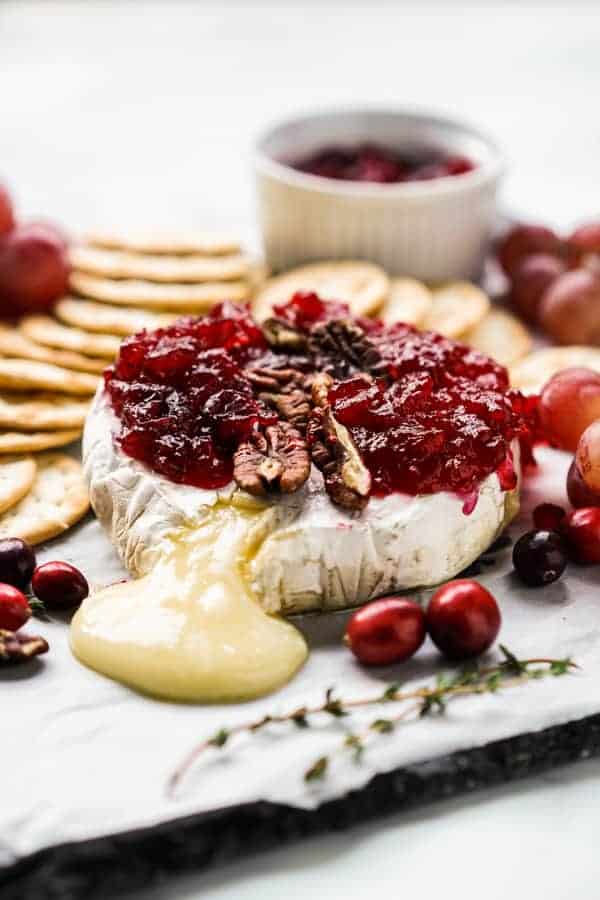 A platter with melted cheese oozing out of baked brie, topped with cranberries and pecans, with crackers surrounding the brie
