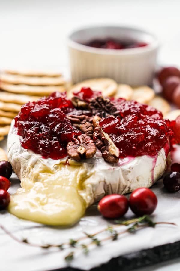 Cheese oozing out of a baked brie that is topped with cranberry sauce and pecans