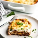 A slice of lasagna with a fork on a plate with a casserole behind it