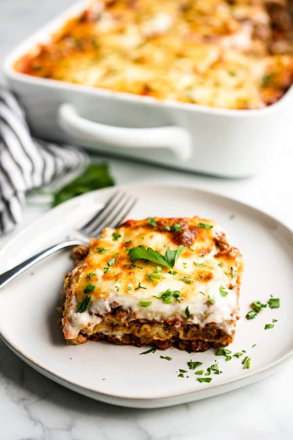 A slice of lasagna with a fork on a plate with a casserole behind it